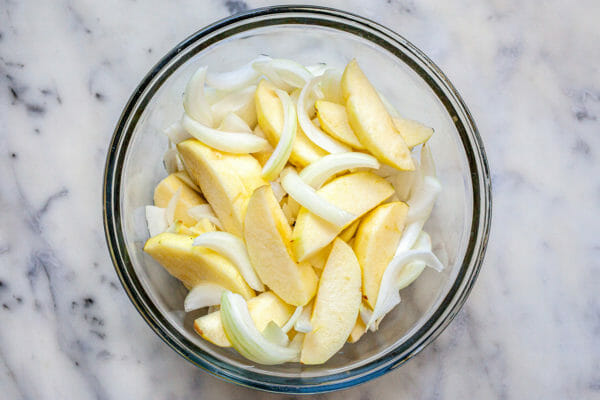  Нарезанные яблоки и лук в стеклянной миске, чтобы приготовить рецепт свиной лопатки. 
